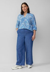 Lichte blouse met een blauw patroon, een v-hals en driekwartmouwen; gecombineerd met losse, donkerblauwe broeken en witte sneakers.