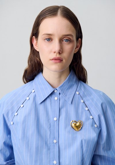 Camicia a righe azzurro chiaro con colletto abbottonato, caratterizzata da bottoni bianchi e un accento di spilla a forma di cuore dorato sul petto.