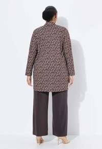 Mujer de espaldas vistiendo un abrigo largo de estampado de leopardo, pantalones anchos oscuros, tacones altos beige y aretes de aro, contra un fondo blanco.