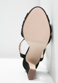 Czarny but na wysokim obcasie z gładkim, teksturowanym wierzchem, regulowanym paskiem na kostkę oraz nagim, karbowanym spodem z niewielkim obcasem.
