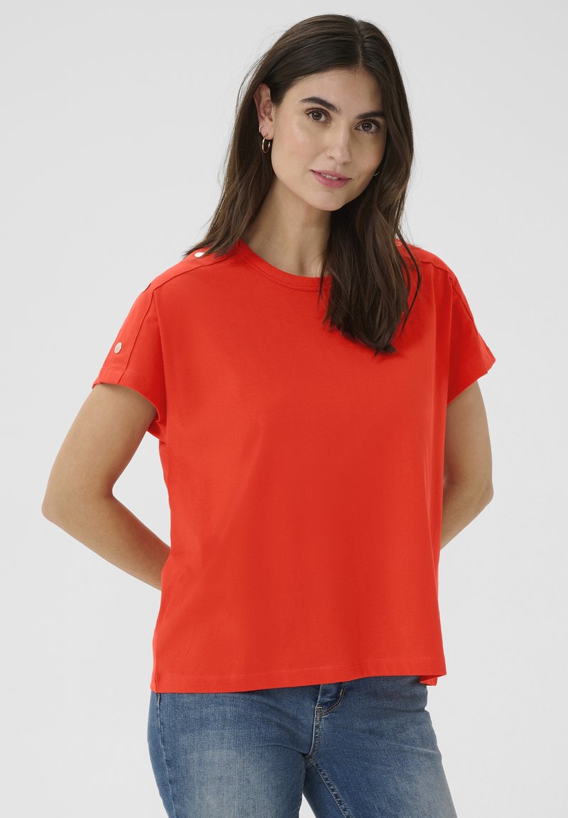 T-shirt rouge à manches courtes en tissu doux, avec des détails de boutons aux épaules et une coupe décontractée, associé à un jean bleu.