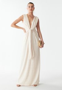 Dolga, svetlobraon obleka z globokim V-izrezom, zavezanem pasom in teksturiranim cvetličnim vzorcem; v paru z zlatim ovojčkom in dodatki.