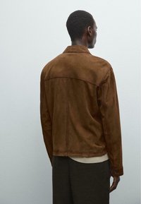 Veste en daim marron avec un col et des coutures piquées, dotée de manches longues et de poignets à boutons, portée sur un haut de couleur claire.
