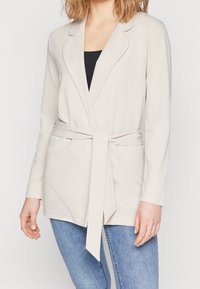 Beige blazerjack met een strikceintuur, lange mouwen en een reverskraag, gemaakt van zachte stof; gedragen over een zwart topje en gecombineerd met jeans.