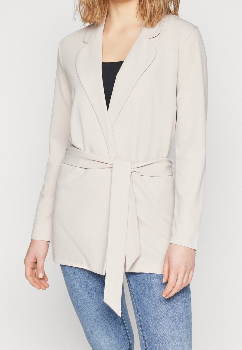 Beige blazerjack met een strikceintuur, lange mouwen en een reverskraag, gemaakt van zachte stof; gedragen over een zwart topje en gecombineerd met jeans.