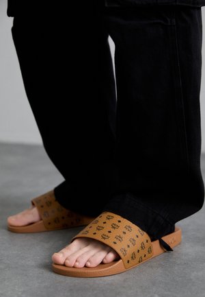 Brown slide sandals with a patterned strap feature a textured footbed and a smooth sole, worn with black pants on a gray surface.