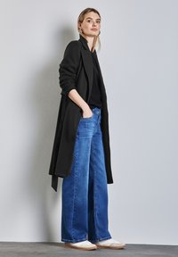 Czarny długi płaszcz na czarnej bluzce, w połączeniu z szerokimi niebieskimi dżinsami i białymi sneakersami. Model stoi z rękami w kieszeniach przy jasnej ścianie.