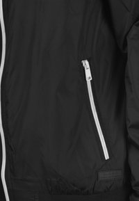 Černá bomber bunda vyrobená z lehkého materiálu, s bílým zipem, elastickými manžetami a texturovanou nášivkou na přední straně.