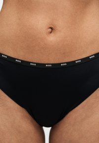 Gros plan sur le bas-ventre d'une personne portant un slip noir avec une ceinture marquée "BOSS" en blanc à plusieurs reprises.