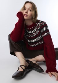 Pull en tricot rouge avec un empiècement à motifs, associé à des ballerines Mary Jane en cuir marron avec une bride et un bout pointu. Le mannequin est assis contre un fond blanc.