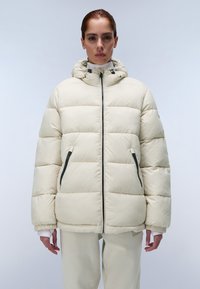 Crème pufferjack met een hoge kraag, horizontale stiknaden, een ritssluiting aan de voorkant en zijzakken die zijnaccentueerd met donkere ritsen; gladde textuur.