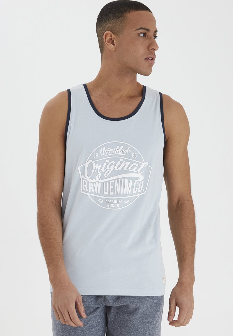 Top cu bretele de un albastru deschis, cu tiv de un navy închis, având un grafic central alb care citește "Original Raw Denim Co." într-un design circular.