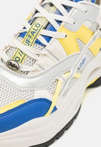 Sneakers Buffalo con tomaia in rete bianca, accenti blu e gialli, sovrapposizioni argentate e lacci multicolore su una suola spessa nera.