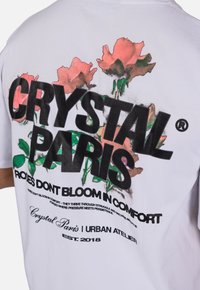 Bílý bavlněný tričko s velkým černým grafickým nápisem "CRYSTAL PARIS" a růžovými květinovými potisky se zelenými akcenty na zadní straně.