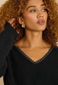 Juodas megztas megztinis su V formos kaklu, puoštas banguotu apvadu ir mažais dekoratyviniais smeigtukais aplink apykaklę. Modelis su garbanotais plaukais.