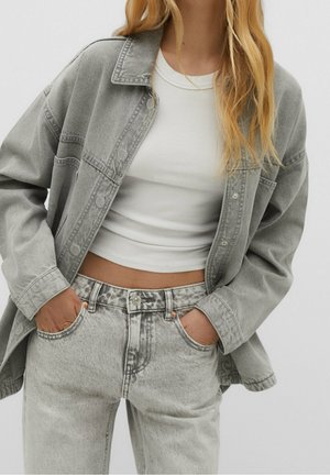 Woman wearing gray oversized denim jacket, white cropped ribbed top, and high-waist light gray jeans with hands in pockets.