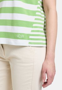 Zielona i biała koszulka w paski z zaokrąglonym dołem, z haftowanym logo 'SCSY' blisko dołu, w połączeniu z beżowymi spodniami.