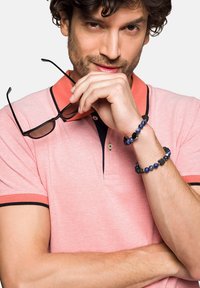 Różowa koszulka polo z pomarańczowym kołnierzykiem i granatowym wykończeniem, zestawiona z ciemnoniebieską bransoletką z koralików. Osoba trzyma okulary przeciwsłoneczne.