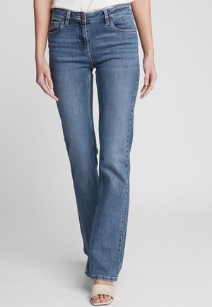 Vrouw in blauwe rechte jeans met een middelhoge taille, knoop- en ritssluiting, en witte open tenen sandalen op een effen achtergrond.