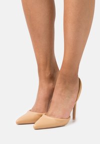 Sapatos de salto alto de bico fino em bege com parte traseira aberta, feitos de couro liso. Apresenta um silhueta elegante e detalhes em costura ao longo das bordas.