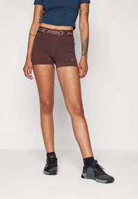 Brune idrettsshorts med en bred, mørk etiket som har teksten "NIKE PRO", laget av elastisk materiale, kombinert med svarte idrettssko.