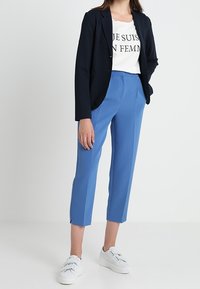 Marineblå blazer, vit T-shirt med svart text och blå korta byxor. Modellen bär vita sneakers. Slät tyg med skräddarsydd passform.