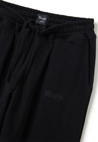 Pantalones deportivos negros hechos de tela suave, con cintura elástica, cordón, bolsillos laterales y un sutil logo "Utopia" bordado en negro.
