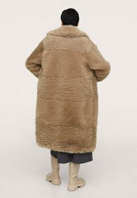 Cappotto in pelliccia sintetica di colore marrone chiaro, design oversize con una morbida texture. Presenta un ampio colletto e dettagli a coste, abbinato a stivali color beige al ginocchio.