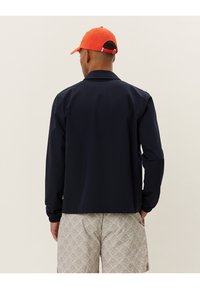 Navy waterproof jacket with long sleeves and elastic cuffs, paired with light patterned shorts and an orange cap. Rear view shows collar detail.