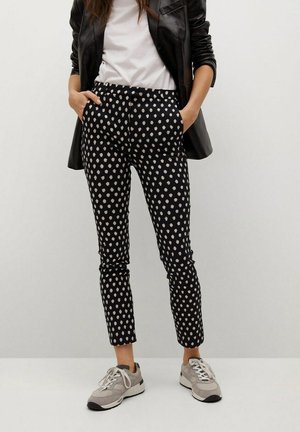 Person trägt schwarz-weiß gepunktete Hose, weißes T-Shirt, schwarze Lederjacke und graue Turnschuhe, steht mit den Händen in den Taschen.