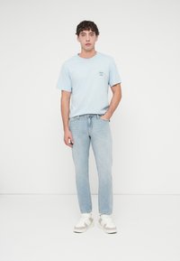 Ανοιχτό μπλε βαμβακερό t-shirt με μικρό τυπωμένο λογότυπο, συνδυασμένο με τζιν παντελόνι ανοιχτού χρώματος και λευκά αθλητικά παπούτσια με μπεζ λεπτομέρειες.