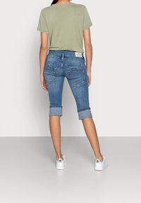 Světle zelené tričko, modré denimové 7/8 kalhoty s ohrnutými nohavicemi, s zadními kapsami a šitým vzorem na zadní straně. Bílé tenisky.