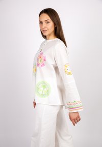 Weißes Leinentop mit langen Ärmeln, ausgestattet mit mehrfarbigen, gestickten runden Mustern und Akzenten entlang der Ärmel. Kombiniert mit weißen Hosen.