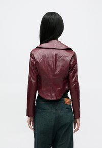 Bordeauxrote Lederjacke in Kurzform mit weitem Kragen und knitteriger Textur, kombiniert mit dunklen Denim-Jeans, die ein Etikett am hinteren Bund tragen.