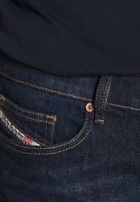 Granatowe dżinsy z żółtym haftem, z przednią kieszenią i widoczną etykietą marki. Obecne są detale w kolorze miedzi, takie jak guziki i nity.