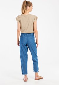 Béžový top s krátkym rukávom v kombinácii s modrými voľnými nohavicami s elastickým pásom. Model nosí hnedé sandále, zobrazené zozadu.