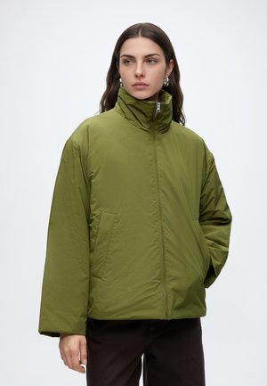 Donna con capelli scuri fino alle spalle che indossa orecchini a cerchio argentati e una giacca imbottita verde oliva con collo alto e tasche anteriori.