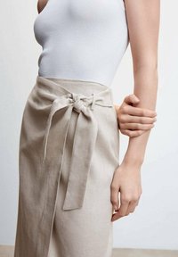 Jupe en lin beige avec un détail de nœud à l'avant et une texture lisse, associée à un débardeur ajusté en maille blanche.