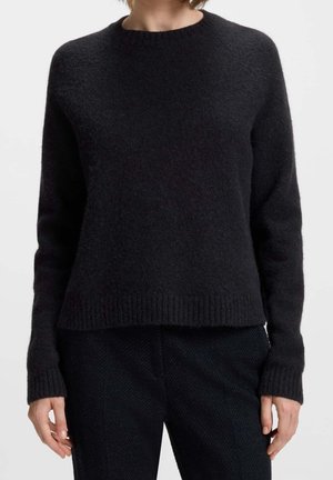 Femme portant un pull en maille noir uni doux avec manches longues et poignets et ourlet côtelés, associé à un pantalon texturé noir.