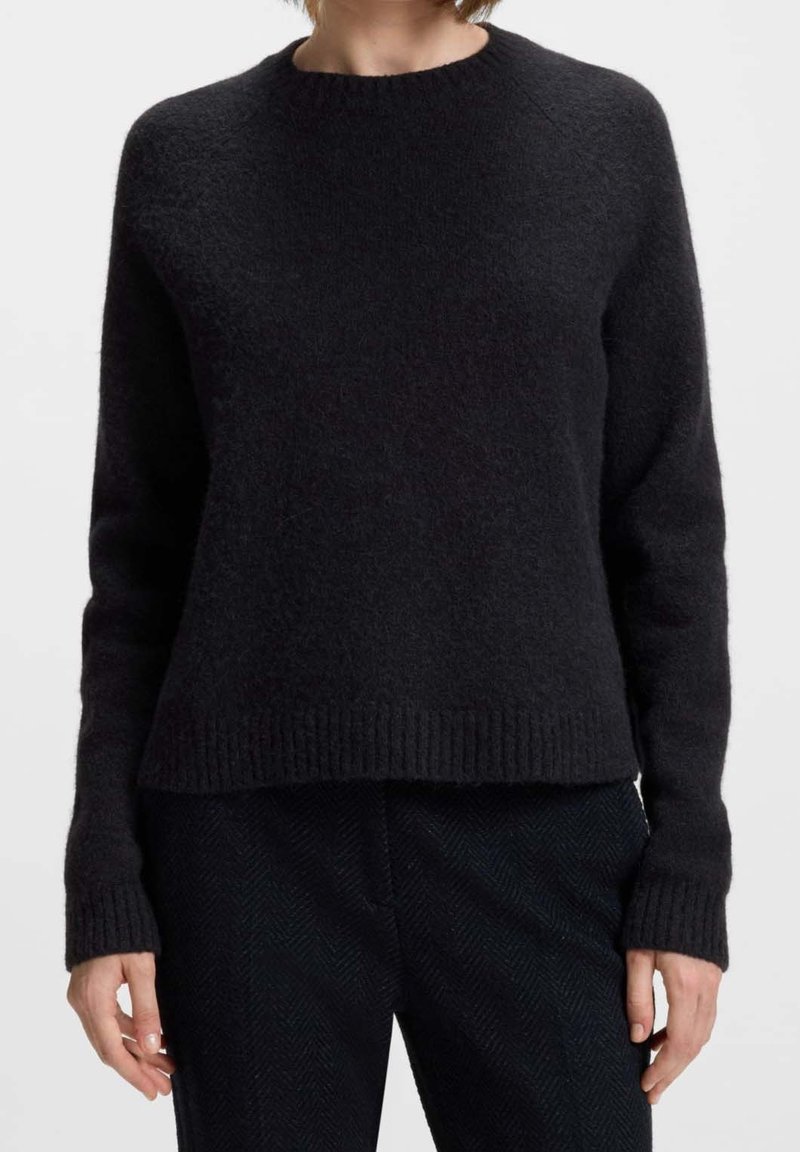 Femme portant un pull en maille noir uni doux avec manches longues et poignets et ourlet côtelés, associé à un pantalon texturé noir.