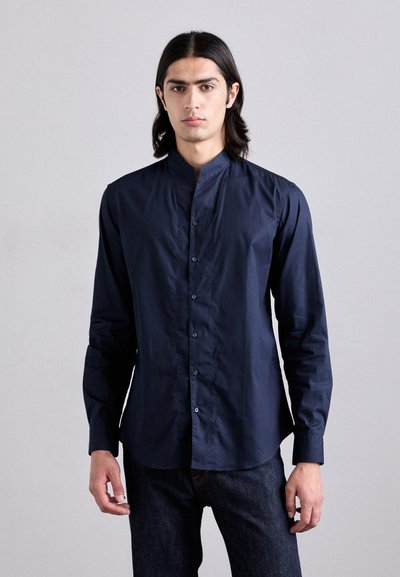 Camicia a maniche lunghe blu navy con colletto alla coreana, chiusura frontale con bottoni e texture liscia. Indossata con jeans in denim scuro.
