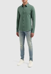 Groene blouse met lange mouwen en kraag, gelegd over lichtblauwe versleten jeans en witte sneakers. Glad stofoppervlak.