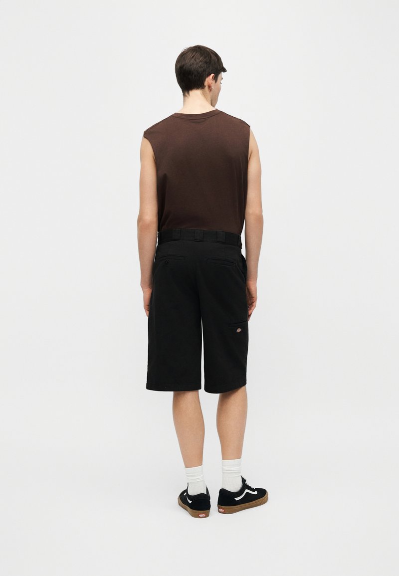 Homme de dos portant un débardeur marron, un short noir jusqu'aux genoux avec ceinture, des chaussettes blanches et des baskets noires avec une bande blanche.