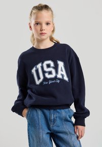 Navy sweatshirt with white "USA" graphic and "New York City" text. Features ribbed neckline and cuffs, paired with light blue baggy jeans.