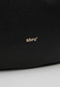 Czarna, teksturowana skórzana powierzchnia z małym, złotym logo marki "abro" umiejscowionym w centrum, blisko dolnej krawędzi.