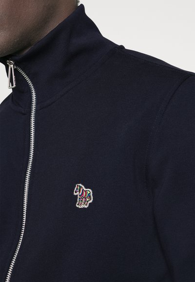Veste zippée navy avec col haut, présentant un logo en broderie colorée de zèbre sur la poitrine gauche et un tissu texturé.