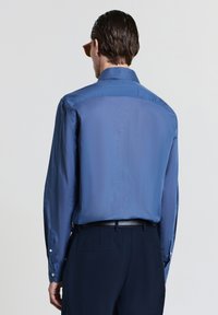 Blaues langärmliges Hemd mit einer glatten Textur, knöpfbaren Manschetten und einem klassischen Kragen, kombiniert mit dunkler Hose. Sichtbare Nähte.