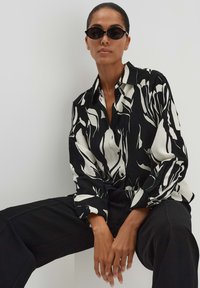 Camicia astratta in bianco e nero con colletto e bottoni, indossata con pantaloni neri. Accessoriata con occhiali da sole neri.