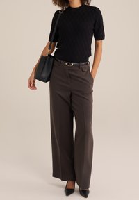 Kvinna som bär en svart kortärmad stickad topp, bruna vida byxor med bälte, svarta spetsiga skor och bär en svart axelväska.