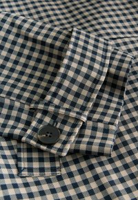 Tmavě modrá a béžová kostkovaná látka zobrazující límec a detail knoflíku. Materiál má hladkou strukturu, se strukturovaným šitím a lesklým knoflíkem.